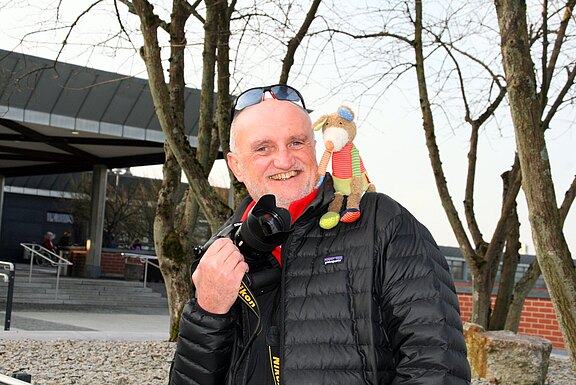 Mann in schwarzer Steppjacke lächelt. Er hält eine Kamera; auf seiner Schulter sitzt ein bunt gekleidetes Stofftier. Aufnahme im Freien vor Bäumen und einem modernen Gebäude.