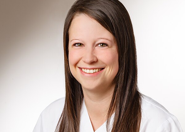 Porträt einer lächelnden jungen Frau mit langem braunem Haar. Sie trägt ein weißes Oberteil ähnlich einem Kittel. Heller, neutraler Hintergrund.