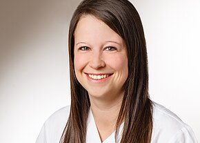 Station 43 Porträt einer lächelnden jungen Frau mit langem braunem Haar. Sie trägt ein weißes Oberteil ähnlich einem Kittel. Heller, neutraler Hintergrund.