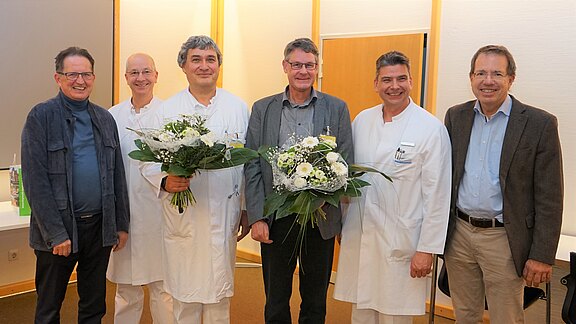 Sechs Personen stehen in einer Gruppe, lächelnd, mit Blumensträußen in den Händen. Einige tragen Arztkittel, andere formelle Kleidung. Der Hintergrund zeigt einen hellen Raum mit neutralen Farben, passend zu einer feierlichen Veranstaltung.