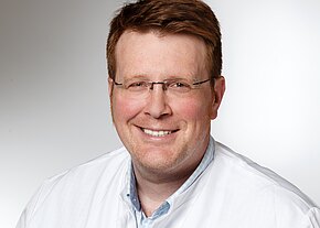 Christian M. Benker Porträt eines lächelnden Mannes mit Brille in weißem Kittel vor neutralem Hintergrund; Kopf-und-Schultern-Aufnahme, direkte Blickrichtung.