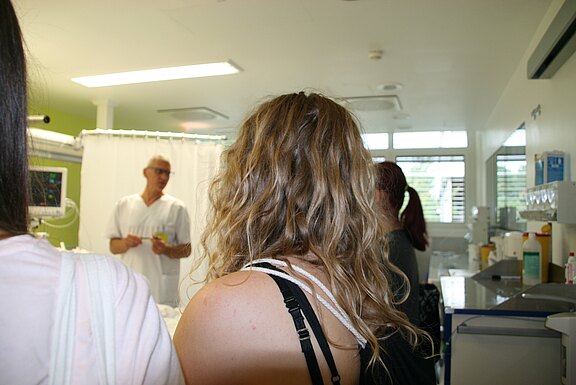 In einem hellen Krankenhauszimmer erklärt medizinisches Personal einer kleinen Gruppe etwas. Die Zuhörer stehen mit dem Rücken zur Kamera; im Hintergrund medizinische Geräte und Vorhang.