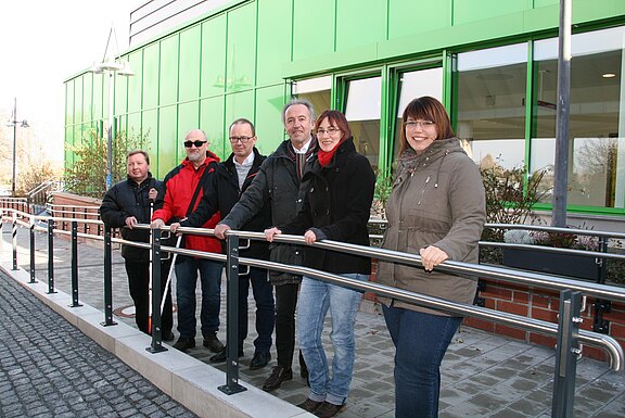 Sechs Erwachsene stehen lächelnd an einem Geländer einer barrierefreien Rampe vor einem modernen, grün verkleideten Gebäude. Sie präsentieren die neue Zugangsrampe und blicken in die Kamera.