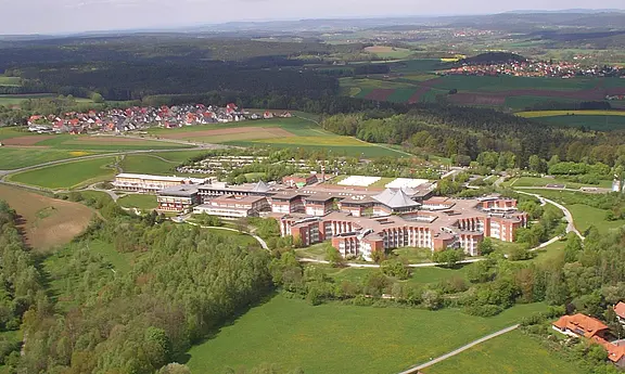 Luftaufnahme eines weitläufigen Gebäudekomplexes mit mehreren Flügeln und modernen Dächern, umgeben von Wiesen, Feldern und Wald. Im Hintergrund verstreute Wohnhäuser und sanfte Hügel. Der Fokus liegt auf dem zentralen Komplex inmitten der grünen Landschaft.