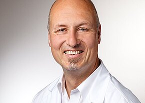 Freundliches Porträt: Ein glatzköpfiger Mann mit kurzem Kinnbart lächelt in die Kamera, trägt ein weißes Oberteil. Kopf-Schulter-Aufnahme vor neutralem Hintergrund, wirkt offen und professionell.