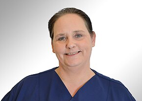 Station 12 B Porträt einer lächelnden Frau mit zurückgebundenen Haaren in dunkelblauer Pflegeuniform. Sie blickt direkt in die Kamera. Heller, neutraler Hintergrund, Fokus auf freundlichem Gesicht und Oberkörper.