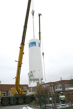 Ein Kran hebt einen großen, zylindrischen Druckbehälter mit der Aufschrift "Linde". Der Behälter wird hochgehoben, während eine Person in Sicherheitskleidung in der Nähe steht, um den Vorgang zu überwachen. Im Hintergrund ist ein Gebäude sichtbar.