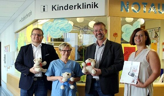 In der Kinderklinik stehen vier Personen zusammen. Sie halten jeweils einen Teddybären in der Hand. Eine Person trägt einen blauen Kittel. Im Hintergrund sind fröhliche Farbdarstellungen sichtbar. Ein Schild über ihnen zeigt 'Kinderklinik' und 'Notaufnahme'.