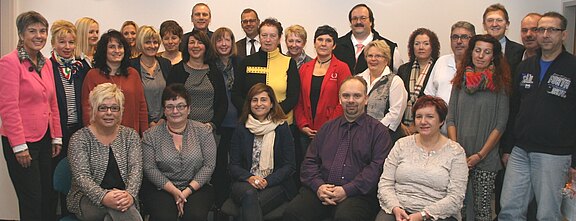 Gruppenfoto von etwa 30 Erwachsenen in einem Raum; vordere Reihe sitzt, hintere Reihen stehen. Die Personen lächeln in die Kamera. Kleidung überwiegend business-casual, gemischte Farben. Neutraler Hintergrund.