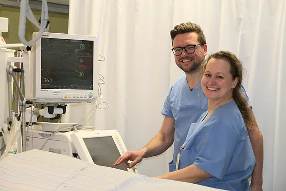Zwei medizinische Fachkräfte in blauer Dienstkleidung stehen neben einem Monitor mit Vitaldaten und bedienen ein Gerät. Sie lächeln; der Raum wirkt wie eine Klinikstation mit Vorhängen. Hauptmotiv: Team arbeitet an Überwachungstechnik am Patientenbett.