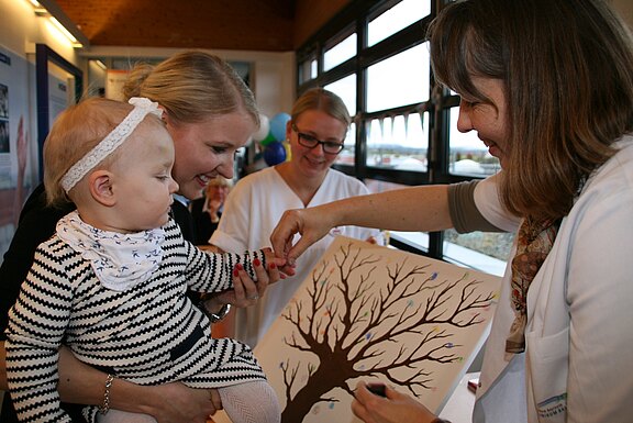 Ein Kleinkind auf dem Arm einer Frau drückt mit Hilfe einer Mitarbeiterin einen eingefärbten Finger auf eine Leinwand mit Baum-Silhouette, um Blätter als Fingerabdrücke zu gestalten. Eine weitere Fachkraft beobachtet. Szene wirkt wie eine kreative Aktivität im Gesundheits- oder Familienzentrum.