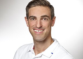 Ein lächelnder Mann mit kurzen, hellen Haaren trägt ein weißes Polohemd. Der Hintergrund ist hell und neutral, was den Fokus auf sein freundliches Gesicht lenkt.