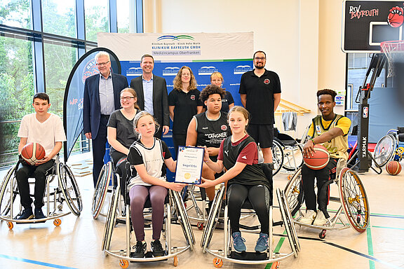 Die Abbildung zeigt eine Gruppe von Jugendlichen im Rollstuhl, die in einer Sporthalle zusammen sind. Einige halten Basketballs, andere präsentieren ein Zertifikat. Im Hintergrund stehen Erwachsene, die die Veranstaltung unterstützen. Die Atmosphäre vermittelt Gemeinschaft und sportlichen Geist.