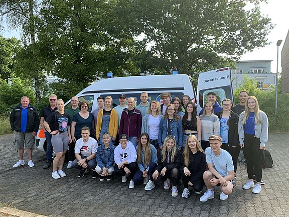 Gruppenfoto einer großen, gemischten Gruppe vor einem weißen Minibus mit Blaulicht im Freien; Personen stehen und hocken in mehreren Reihen und lächeln in die Kamera.