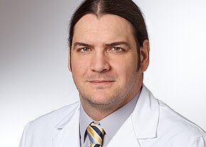 Porträt eines Mannes mit zurückgebundenen dunklen Haaren. Er trägt einen weißen Kittel, graues Hemd und gestreifte Krawatte. Neutrale Mimik, Blick in die Kamera, heller Hintergrund. Wirkt wie ein medizinischer Fachmann.