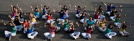 Eine Gruppe von etwa 30 Menschen sitzt auf dem Boden, trägt bunte Oberteile und weiße Hosen. Sie lächeln, heben die Arme und wirken fröhlich. Die Pose vermittelt ein Gefühl von Gemeinschaft und Freude.