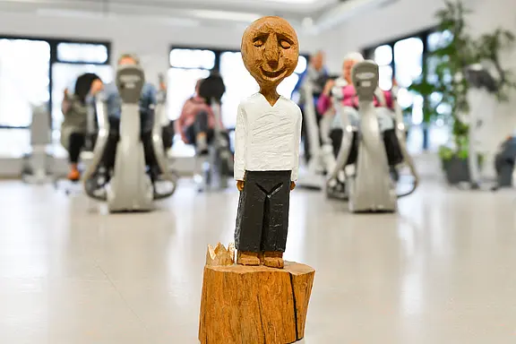 Im Vordergrund steht eine kleine, lächelnde Holzfigur mit weißem Oberteil und dunkler Hose auf einem Holzsockel. Im unscharfen Hintergrund trainieren mehrere Menschen auf Heimtrainern in einem hellen Raum.