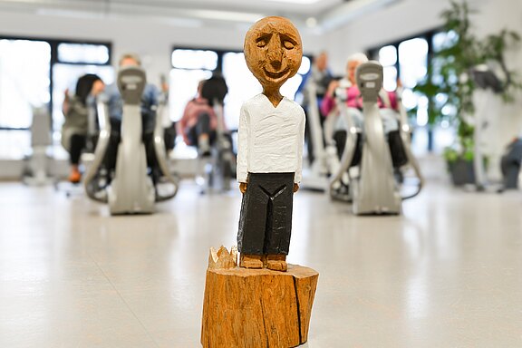 Im Vordergrund steht eine kleine, lächelnde Holzfigur mit weißem Oberteil und dunkler Hose auf einem Holzsockel. Im unscharfen Hintergrund trainieren mehrere Menschen auf Heimtrainern in einem hellen Raum.