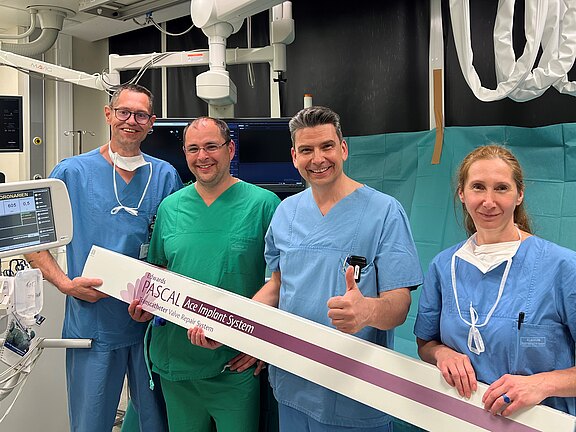 Vier OP-Mitarbeitende in grüner und blauer Kleidung posieren im Klinikraum und halten gemeinsam eine lange Verpackung mit der Aufschrift „PASCAL Ace Implant System“; einer zeigt den Daumen hoch. Die Szene vermittelt die Vorstellung einer erfolgreichen medizinischen Prozedur oder neuen Ausrüstung.