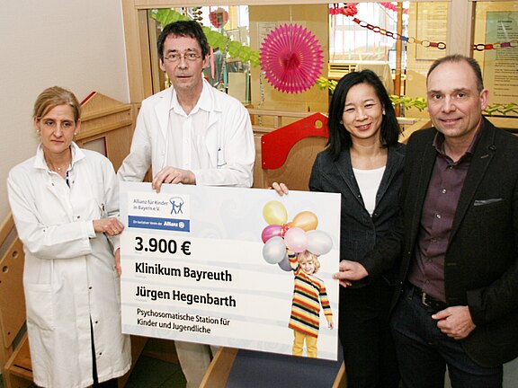 Eine Gruppe von vier Personen hält einen Scheck über 3.900 Euro. Der Scheck ist für das Klinikum Bayreuth und den Projektleiter Jürgen Hegenbarth, zur Unterstützung der psychosomatischen Station für Kinder und Jugendliche. Im Hintergrund sind kindgerechte Elemente sichtbar.