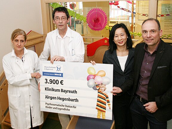 Eine Gruppe von vier Personen hält einen Scheck über 3.900 Euro. Der Scheck ist für das Klinikum Bayreuth und den Projektleiter Jürgen Hegenbarth, zur Unterstützung der psychosomatischen Station für Kinder und Jugendliche. Im Hintergrund sind kindgerechte Elemente sichtbar.