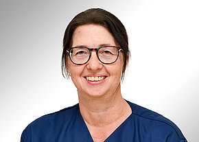 Station 12 A Porträt einer lächelnden Frau mit Brille in dunkelblauer Arbeits- bzw. Pflegekleidung vor neutralem Hintergrund.
