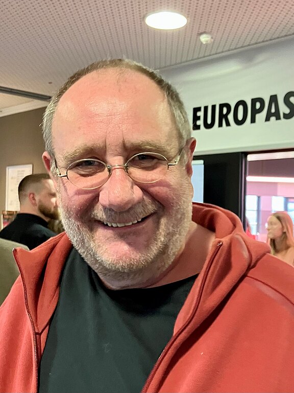 Porträt eines lächelnden, mittelalten Mannes mit kurz geschnittenem Haar, Brille und Bartstoppeln, in roter Kapuzenjacke über schwarzem T‑Shirt, aufgenommen in einem Innenraum.