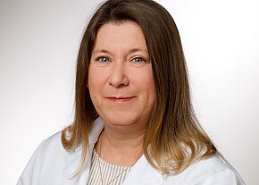 Ein Portrait einer Frau mit langen, glatten Haaren, die in einem weißen Laborkittel gekleidet ist. Sie lächelt leicht und schaut direkt in die Kamera. Der Hintergrund ist hell und neutral.