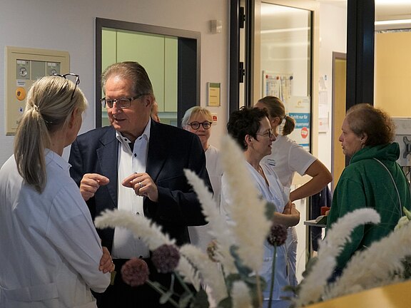 In einem Klinikflur sprechen mehrere Pflegekräfte und Ärztinnen in weißer Kleidung mit einem Mann im Anzug; daneben steht eine Person in grünem Hoodie. Die Gruppe wirkt im Gespräch über medizinische Belange; Klinikschilder und Geräte im Hintergrund.