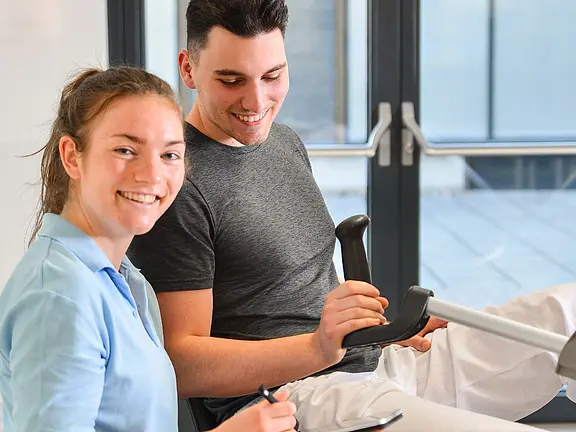 Physiotherapeutin begleitet einen jungen Mann beim Training an einem Ergometer; beide lächeln. Die Therapeutin notiert Werte auf einem Klemmbrett, der Patient hält den Griff des Geräts. Szene vermittelt Reha oder gesundheitsorientiertes Training in heller Umgebung.