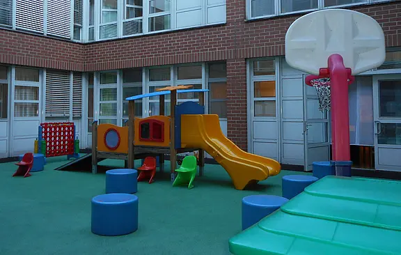 Kinderspielbereich in einem Innenhof: buntes Klettergerüst mit Rutsche, kleine Plastikstühle und Polsterpodeste auf weichem Boden. Rechts ein Basketballkorb, daneben große Schaumstoffmatten. Umgebung wirkt sicher und für Vorschulkinder geeignet.