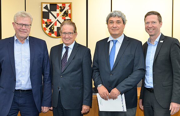 Vier Männer in Anzügen stehen nebeneinander für ein offizielles Foto in einem Sitzungssaal. Einer hält ein Dokument. Im Hintergrund ist ein Wappen an der Wand. Neutrale, freundliche Gesichter.