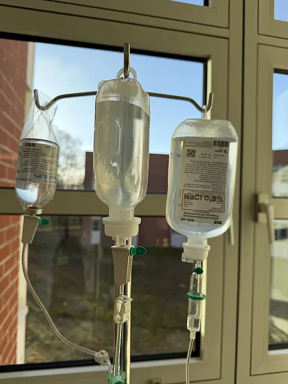 Drei Infusionsbeutel hängen an einem Ständer vor einem Fenster; klare Flüssigkeiten, einer sichtbar mit NaCl 0,9 % beschriftet. Schläuche mit Tropfkammern führen nach unten. Klinische Umgebung bei Tageslicht.