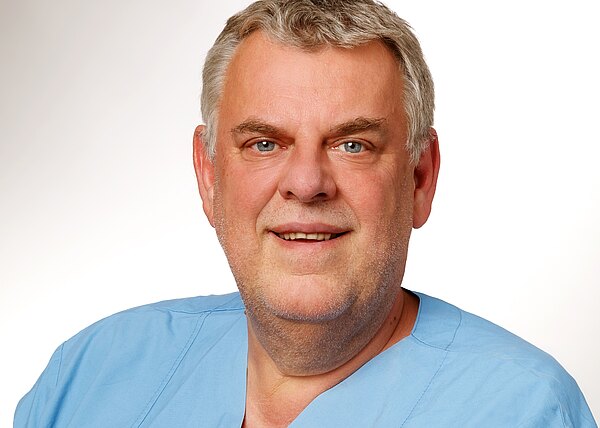 Porträt eines lächelnden Mannes mittleren Alters in hellblauer medizinischer Kleidung vor neutralem Hintergrund; Kopf und Schultern frontal sichtbar.