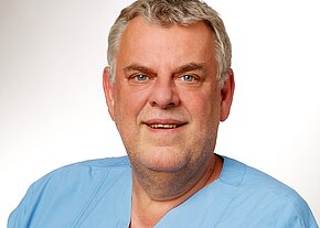 Station 58 Porträt eines lächelnden Mannes mittleren Alters in hellblauer medizinischer Kleidung vor neutralem Hintergrund; Kopf und Schultern frontal sichtbar.