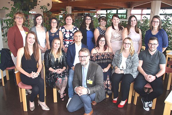Gruppenfoto in einem Innenraum: etwa zwanzig junge Erwachsene und zwei ältere Personen posieren lächelnd, teils sitzend, teils stehend. Kleidung überwiegend festlich bis leger. Es wirkt wie eine Abschluss- oder Teamaufnahme bei einer Veranstaltung.