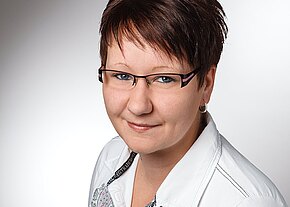 Michaela Kretz Porträtfoto: lächelnde Frau mit kurzer brauner Frisur und rechteckiger Brille, trägt eine weiße Bluse. Halbprofil vor neutralem, hellgrauem Hintergrund.