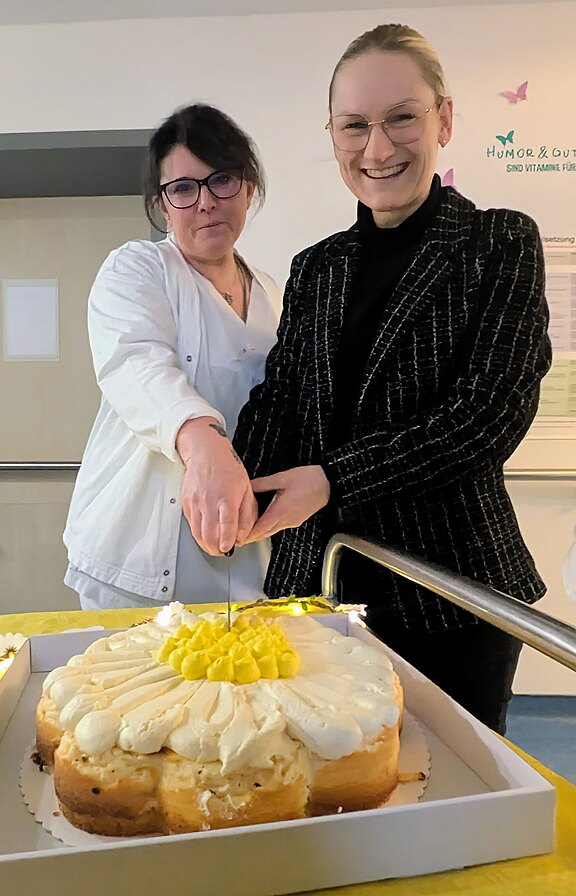 Zwei Frauen schneiden gemeinsam eine große Sahnetorte mit gelber Creme an, lächelnd in einem Innenraum. Die Torte steht in einem Karton auf dem Tisch; beide halten zusammen das Messer.