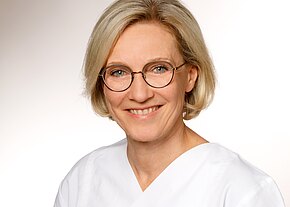 Notaufnahme Kinderklinik Porträt einer lächelnden Frau mittleren Alters mit kurzem blondem Haar und runder Brille, in weißer medizinischer Kleidung vor hellem, neutralem Hintergrund.