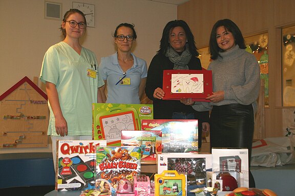 Vier Frauen, zwei in Pflegekleidung, stehen in einer Kinderklinik hinter einem Tisch voller Spielzeuge und Spiele. Zwei halten ein Kinderbild hoch. Die Szene wirkt wie eine Spendenübergabe für junge Patientinnen und Patienten.