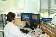 Person im weißen Laborkittel mit Headset sitzt am Schreibtisch und arbeitet an zwei Monitoren. Auf den Bildschirmen sind Programme oder Daten zu sehen, Labor-/Büroumgebung.
