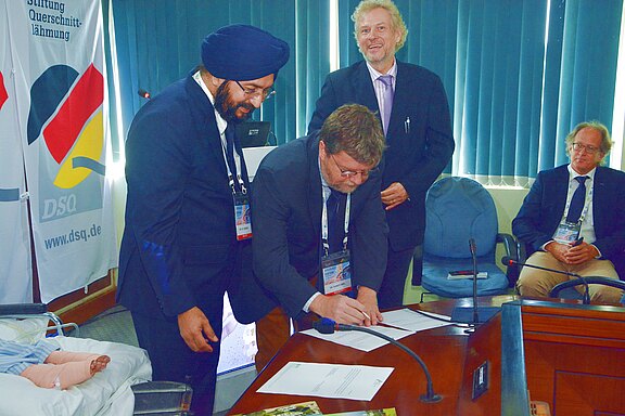 Mehrere Männer in Anzügen bei einer offiziellen Unterzeichnung: Einer beugt sich über einen Tisch und signiert Dokumente, ein anderer weist darauf, weitere Teilnehmer beobachten im Konferenzraum. Mikrofone, Namensschilder und Flaggen deuten auf eine formelle Vereinbarung oder Partnerschaft hin.