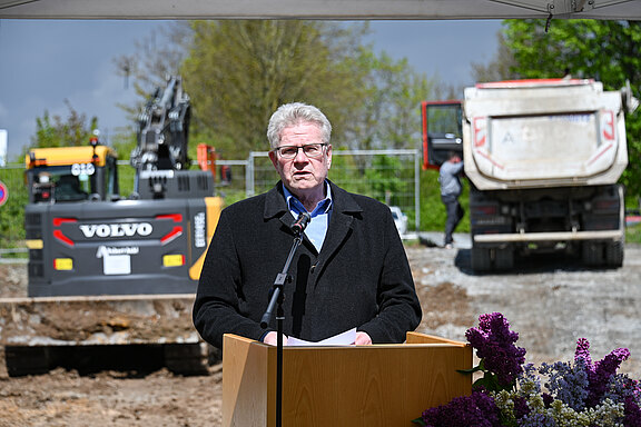 Ein Mann hält eine Rede am Podium auf einer Baustelle. Er spricht ins Mikrofon und hält Papiere. Hinter ihm stehen ein Bagger und ein Lastwagen. Die Szene wirkt wie ein Spatenstich oder Projektstart.