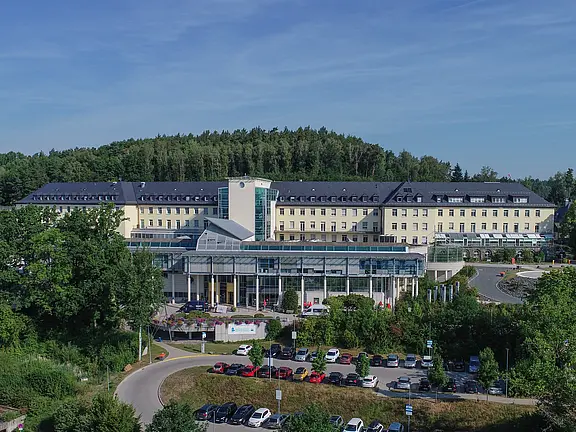 Luftaufnahme eines großen Klinikkomplexes: modernes, verglastes Eingangsgebäude vor langem, mehrstöckigem Haupttrakt. Davor Parkplätze und geschwungene Zufahrt; dahinter bewaldeter Hügel.