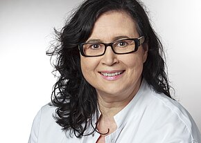 Station 11 Porträt einer lächelnden Person mit dunklen, lockigen Haaren und schwarzer Brille, in weißem Kittel, vor neutralem Hintergrund.