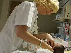 Eine Krankenschwester kümmert sich um ein Säugling, während sie ihn auf einer Liege hält. Sie überprüft das Baby, das in einem hellen Strampler gekleidet ist. Die Atmosphäre wirkt fürsorglich und gemütlich.