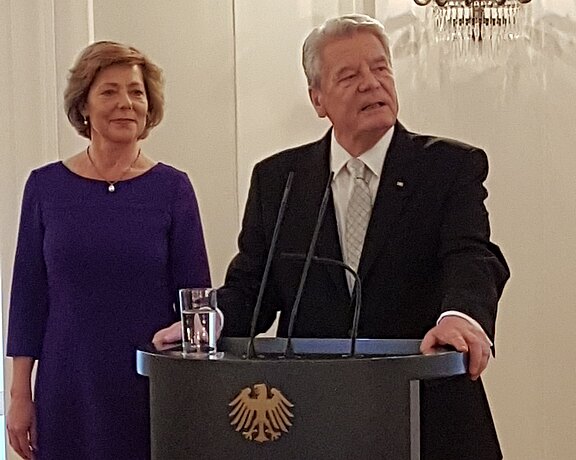 Zwei Personen stehen vor einem Rednerpult mit dem deutschen Bundesadler. Ein Mann in einem schwarzen Anzug spricht, während eine Frau in einem lila Kleid neben ihm steht und aufmerksam zuhört. Der Kontrast zwischen ihrer Kleidung und dem neutralen Hintergrund betont die Szene.