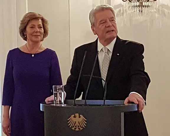 Zwei Personen stehen vor einem Rednerpult mit dem deutschen Bundesadler. Ein Mann in einem schwarzen Anzug spricht, während eine Frau in einem lila Kleid neben ihm steht und aufmerksam zuhört. Der Kontrast zwischen ihrer Kleidung und dem neutralen Hintergrund betont die Szene.