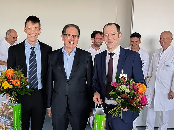 Drei Männer in Anzügen stehen im Vordergrund, jeder mit einem Strauß Blumen in der Hand. Sie lächeln freundlich. Im Hintergrund sind mehrere Personen in weißen Kitteln sichtbar, die ebenfalls lächeln. Die Szene vermittelt eine festliche und herzliche Atmosphäre.