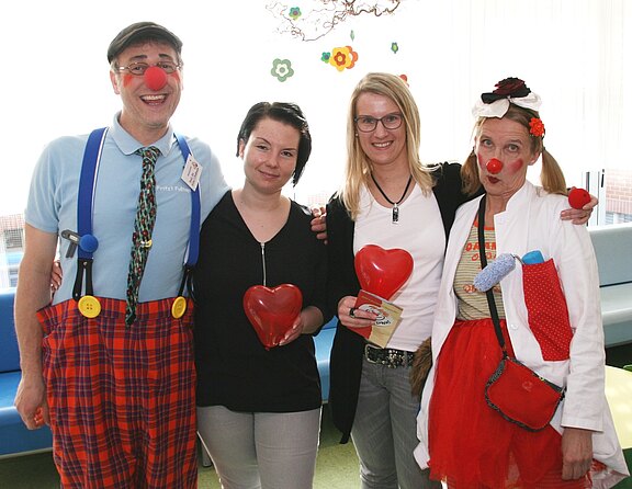 Vier Erwachsene stehen zusammen und lächeln. Zwei als Klinikclowns verkleidet mit roten Nasen und bunter Kleidung, zwei in Alltagskleidung. Drei halten rote herzförmige Objekte. Stimmung freundlich, Unterstützungs- oder Spendenaktion angedeutet.